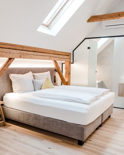 Ein Doppelbett das von der Seite fotografiert wurde und vom Dachfenster beleuchtet wird, während man die Schiebetür zum Badezimmer im Hintergrund erkennen kann