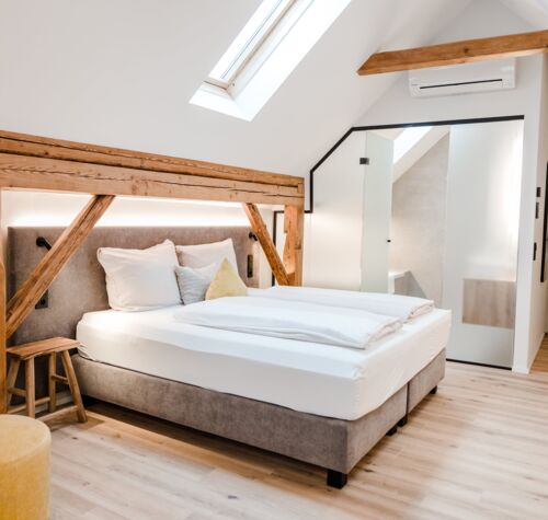 Ein Doppelbett das von der Seite fotografiert wurde und vom Dachfenster beleuchtet wird, während man die Schiebetür zum Badezimmer im Hintergrund erkennen kann