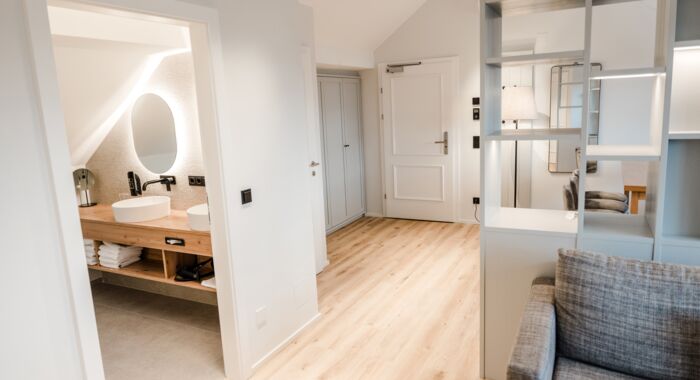 Ein Großraum Bild von dem Familienzimmer wo auf der rechten Seite die Couch und ein Schrank dahinter zusehen ist und auf der anderen Seite sieht man in das Badezimmer hinein.