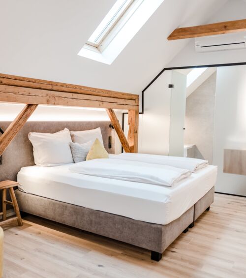 Ein Doppelbett das von der Seite fotografiert wurde und vom Dachfenster beleuchtet wird, während man die Schiebetür zum Badezimmer im Hintergrund erkennen kann